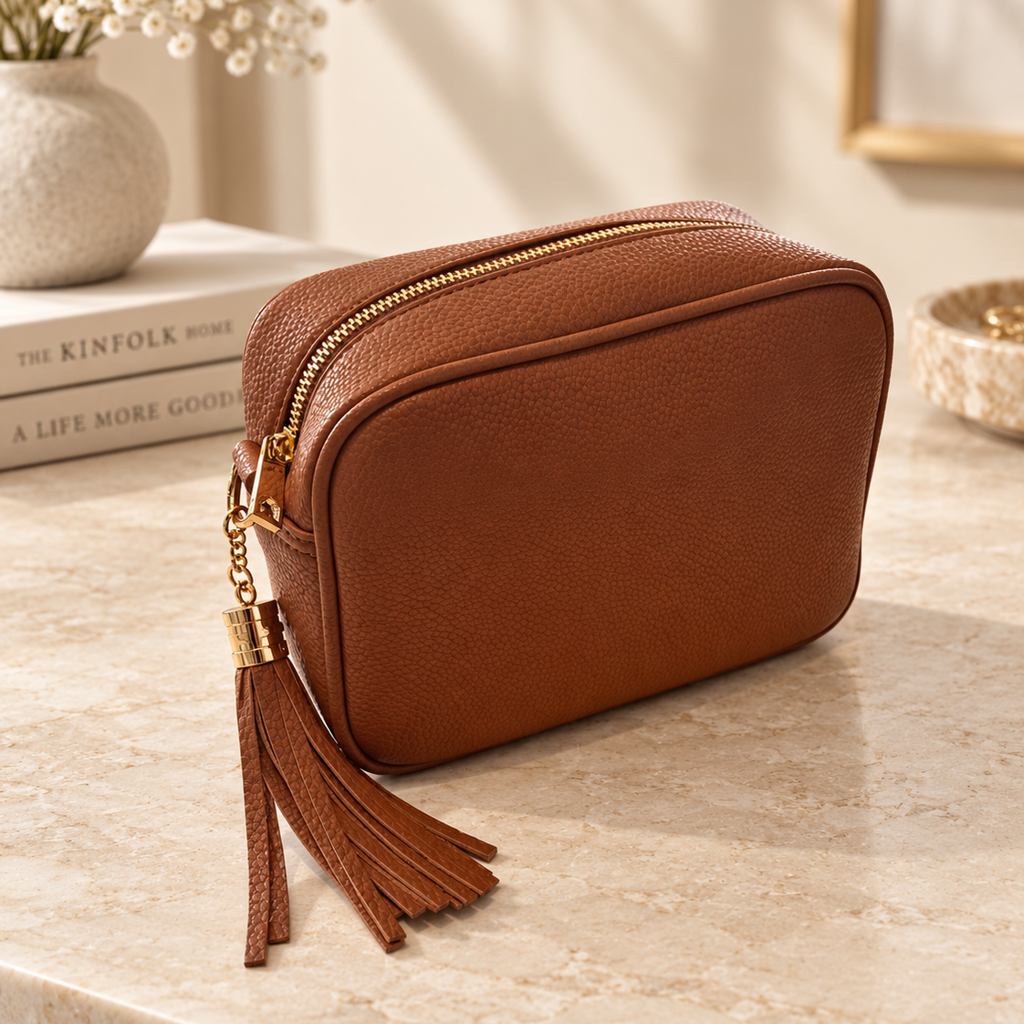 A brown pebbled faux-leather cosmetic pouch with a gold zipper and a matching tassel charm, resting on a beige marble surface in a styled interior setting.