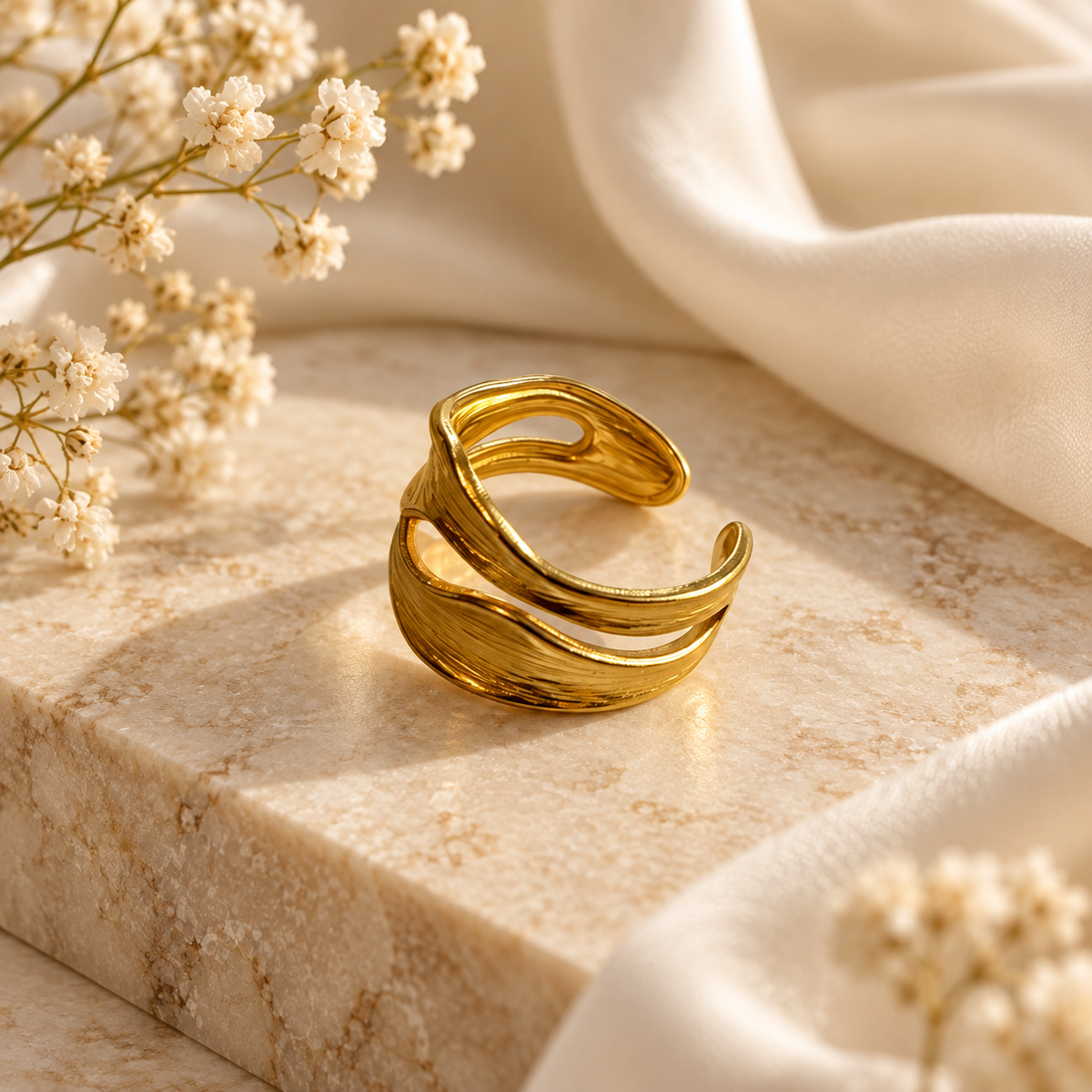 A gold-toned open ring with a textured, flowing layered design, displayed on a beige marble block with white fabric and dried baby's breath flowers.