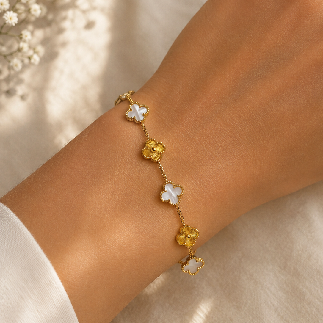 A delicate gold chain bracelet with alternating white and yellow flower charms, shown worn on a person's wrist against a soft beige fabric background.