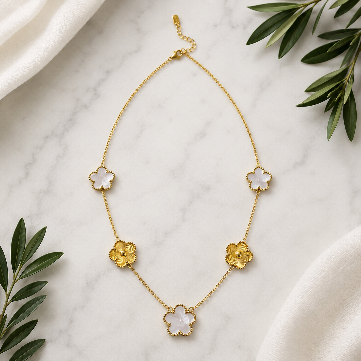 A gold chain necklace with five alternating gold and white mother-of-pearl four-petal flower charms, displayed on a white marble surface with green leaves and draped white fabric.