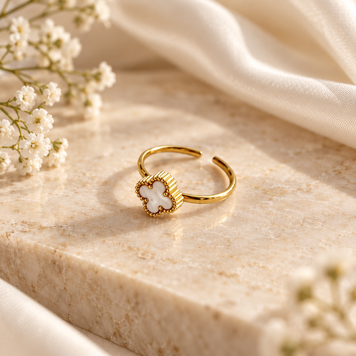 A gold adjustable ring with a white clover-shaped inlay, styled on a beige marble block with white fabric and baby's breath flowers.